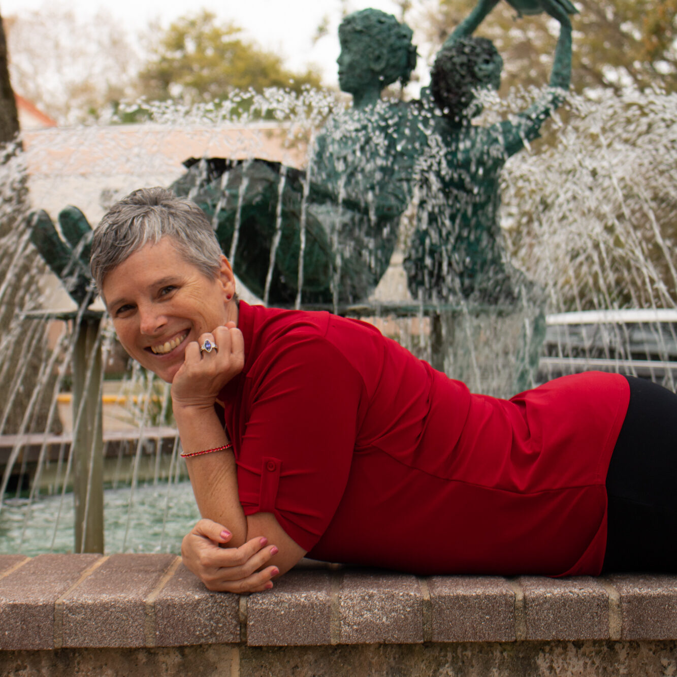Amy Levesque laying playfully in front of water fountain. Smiling expression, red polo